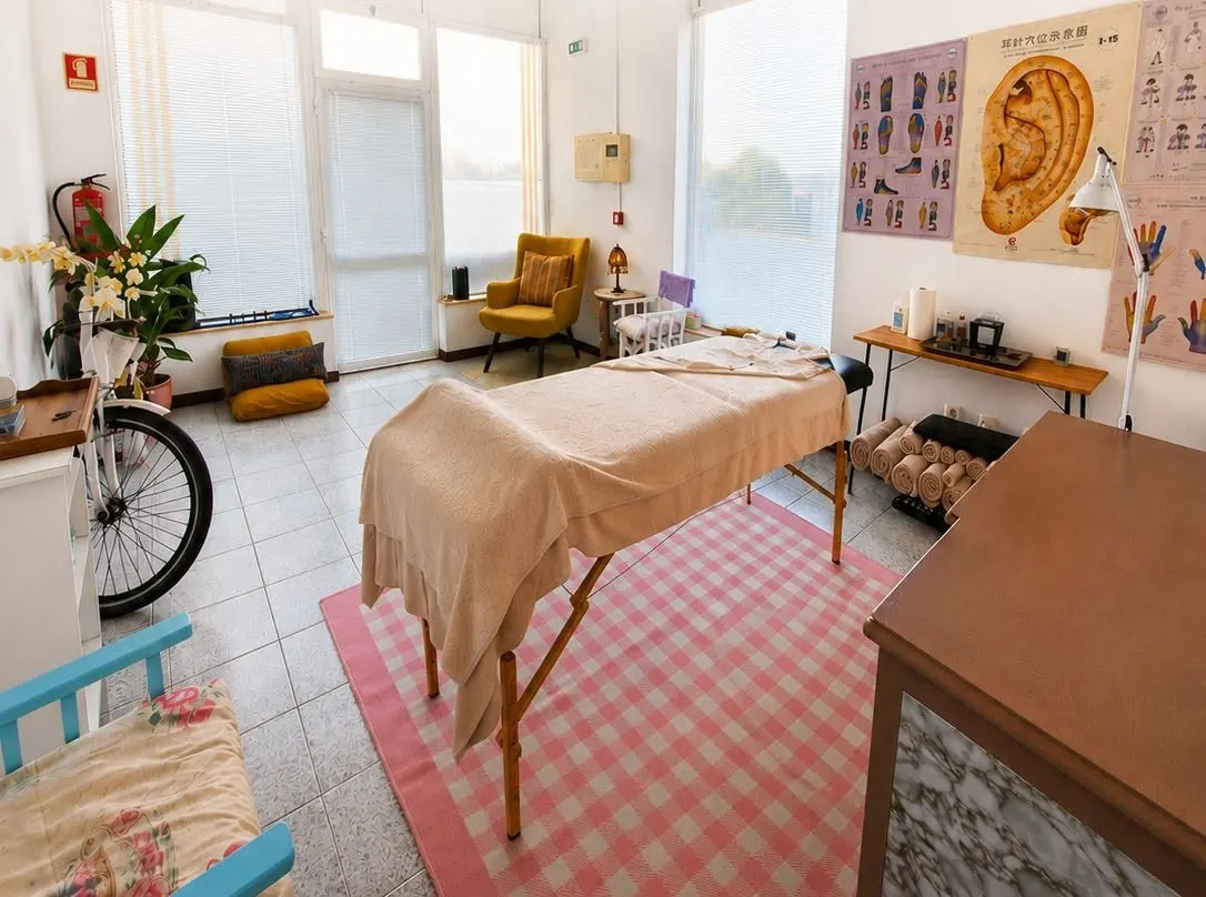 Sala de terapias da Rainbow Tree em Vila Nova de Poiares: marquesa de massagem, tapete aos quadrados rosa, poltrona amarela, plantas e mapas de reflexologia e auriculoterapia na parede